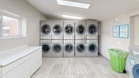 Located at 1 Zachary Ln in Hamilton, NJ, this shared laundry facility is a well-organized, single-floor space designed to efficiently serve its users. The room features a clean, modern aesthetic with light gray large-format tile flooring, white walls, and a neutral color palette that creates a bright and welcoming environment.

The facility is equipped with a generous number of front-loading washing machines arranged along multiple walls, providing ample capacity for high-demand use. A bank of stacked front-load dryers lines one wall, maximizing the available floor space. A white countertop cabinet along one wall offers a convenient folding and sorting surface. A utility sink is also present, adding practical functionality to the space.

Natural light enters through multiple windows and skylights, supplemented by recessed fluorescent ceiling lighting that ensures consistent illumination throughout. The room is climate-controlled via a wall-mounted HVAC unit. Two interior doors provide access to adjacent areas, and a clearly marked exit door with direct outdoor access ensures easy egress.

Waste management is addressed with a dedicated recycling bin on-site. Signage indicates that the machines support mobile payment, offering a modern and convenient payment option for users. Overall, the facility is thoughtfully laid out, clean, and fully equipped to meet the everyday laundry needs of residents.