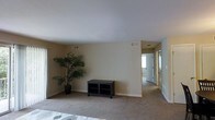 This single-floor residence offers a clean, understated design characterized by neutral beige walls, smooth ceilings, and wall-to-wall carpeting that lends a comfortable, cohesive feel throughout. The main living area is spacious and open, providing ample room for flexible furniture arrangements. Multiple HVAC vents are positioned thoughtfully across the ceiling, ensuring consistent climate control. A closet with sliding doors is integrated into the living space, offering convenient built-in storage. The room opens naturally into a hallway that connects to additional areas of the home, including what appears to be a bedroom with a bright window that draws in natural light. The overall color palette—consisting of soft, warm neutrals—creates a calm and welcoming ambiance. White baseboards provide a clean contrast against the carpeted floors, adding a subtle architectural detail that ties the space together. The layout is straightforward and functional, making it well-suited for a variety of living arrangements. The combination of practical features, including generous storage, good ventilation, and a bright, airy feel, makes this a versatile and comfortable single-floor living space.