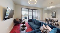 This contemporary single-floor residence showcases a thoughtfully designed open-plan layout that seamlessly integrates the living, dining, and kitchen areas into one cohesive space. Light-toned hardwood flooring runs throughout, complementing the clean white walls and creating a bright, airy atmosphere.

The living area is a standout, anchored by a plush tufted teal sofa, a vibrant patterned area rug, and a striking circular halo pendant light overhead. Floor-to-ceiling sliding glass doors flood the room with natural light and open directly onto a private balcony, where outdoor seating captures sweeping views of the surrounding neighborhood. A wall-mounted flat-screen television and a sleek accent chair complete the space.

The kitchenette is efficiently designed with white cabinetry, a grey stone countertop, and a stainless steel sink. Open timber shelving adds a warm, modern touch, while an under-counter refrigerator and built-in microwave provide full functionality. A compact dining table with black bar stools serves as a versatile eat-in area adjacent to the kitchen.

A wall-mounted split-system air conditioning unit ensures year-round comfort. The overall ambiance is polished and contemporary, balancing functional design with curated, stylish furnishings throughout.