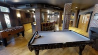 This dynamic entertainment space is designed for socializing and recreation, combining industrial-chic aesthetics with a warm, inviting atmosphere. The layout is anchored by a full-size billiards table positioned prominently on rich hardwood flooring, making it a natural focal point of the room. Structural brick columns rise throughout the space, adding a bold architectural character that complements the exposed ductwork overhead, which runs along a curved, dark-finished ceiling to reinforce the industrial style. A well-appointed bar area takes center stage in the background, featuring a sleek countertop and a row of classic red upholstered bar stools, ideal for gathering and conversation. Pendant lighting and wall sconces provide warm, layered illumination throughout. Additional cabinetry with yellow accent finishes lines one wall, offering both storage and visual interest. A foosball or gaming table is also visible, further enhancing the recreational offerings of the space. Colorful wall art provides a vibrant finishing touch near the bar area. The open-plan design ensures easy flow between the various activity zones, making this a well-rounded entertainment destination suited for hosting and leisure.