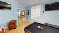 This single-floor community amenity space is thoughtfully designed for recreation and social gathering, offering a well-appointed layout with consistent finishes throughout. The main game room serves as the centerpiece, featuring a full-size pool table set against light blue walls, warm hardwood flooring, and crisp white crown molding. A wall-mounted flat-screen television and comfortable leather seating chairs make this area ideal for both active play and relaxed viewing. Decorative framed artwork adds a personalized touch to the walls. The game room connects seamlessly to a lounge and living area through elegant glass-paneled French doors, which allow natural light to flow freely between spaces. The adjacent living area continues the warm hardwood flooring and is filled with natural light from large windows dressed with horizontal blinds. A cozy bistro-style nook, accented with a deep navy accent wall and a small high-top table with bar-height seating, offers an intimate spot for casual conversation. The space also includes access to a water fountain alcove and direct exterior access through glass-paned exit doors, providing views of surrounding greenery. The overall ambiance is bright, welcoming, and versatile, combining recreational and social functions in a cohesive, well-maintained environment.
