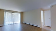 This single-floor space offers a clean, functional layout designed for comfortable everyday living. The main living area is highlighted by warm hardwood flooring that extends throughout, lending a sense of continuity and warmth to the interior. A large sliding glass door fitted with vertical blinds serves as a prominent architectural feature, allowing an abundance of natural light to fill the room while also providing direct access to an outdoor area. The neutral, light-toned walls contribute to a bright and airy ambiance, making the space feel open and welcoming. Baseboard heating units run along the lower perimeter of the living area, ensuring efficient and even climate control throughout the colder months. The smooth, white ceiling complements the understated color palette, enhancing the overall sense of openness. A doorway visible from the main living area suggests additional rooms beyond, offering further functional space within the unit. The overall design is straightforward and practical, with a neutral aesthetic that provides a versatile backdrop suited to a variety of interior styles and furnishings.