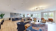 Located at 7810 Contee Rd in Laurel, Maryland, this thoughtfully designed community lounge space offers a vibrant and welcoming atmosphere across a single, generously sized floor. The open-concept layout is anchored by light hardwood flooring that runs throughout, complementing the clean white walls and modern finishes.

The main lounge area features a curated mix of seating, including plush tufted sofas, velvet accent chairs in deep navy blue, and leather lounge chairs arranged around a bold, multicolored area rug. An eye-catching branching brass chandelier serves as a focal point overhead. A wall-mounted flat-screen TV is set into a textured dark tile accent wall, adding depth and visual interest.

A built-in bar area with patterned tile backsplash, open shelving, and counter seating provides a stylish social hub. Adjacent to this is a dining nook outfitted with booth-style banquette seating, pedestal tables, and a striking red pendant light, ideal for casual gatherings.

Large windows on multiple walls flood the space with natural light and offer views of the surrounding greenery. Curated artwork and neon signage add personality throughout. An entry corridor with built-in bench seating and textured accent walls leads to double glass exit doors, rounding out this well-appointed communal retreat.
