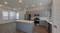 This contemporary single-floor residence showcases a clean, modern aesthetic throughout, highlighted by a bright, neutral color palette and thoughtful finishes. The heart of the home is a well-designed kitchen featuring sleek gray shaker-style cabinetry extending to the ceiling for generous storage, a white quartz countertop island with built-in outlets, and a full suite of stainless steel appliances including a gas range, microwave, French door refrigerator, and dishwasher. White subway tile backsplash and light wood-look luxury vinyl plank flooring complete the contemporary look. The open layout connects the kitchen seamlessly to adjacent living areas, with a glass-panel entry door that fills the space with natural light. The bedrooms offer a calm retreat, with some featuring soft carpeting, ceiling fans, and windows dressed with horizontal blinds. One bedroom showcases the same wood-look flooring as the main living areas. The bathroom is a standout, finished in elegant marble-look tile from floor to ceiling. It includes a gray vanity with white quartz countertop, a undermount sink with brushed gold fixtures, and a frameless glass-enclosed walk-in shower with marble-tiled walls and a built-in niche. Recessed lighting throughout the home ensures a bright, inviting atmosphere in every room.