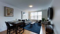 This single-floor residence offers a thoughtfully designed open-concept layout that seamlessly integrates the living and dining areas into one cohesive, functional space. Warm hardwood flooring runs throughout, providing a rich foundation that complements the clean, neutral color palette of white walls and modern finishes.

The living area is anchored by a large area rug in deep navy, adding a bold contrast to the light tones of the room. A comfortable gray sofa and a sleek accent chair are arranged to maximize conversation and relaxation, while a contemporary coffee table ties the seating arrangement together. A large window along the exterior wall allows abundant natural light to pour into the space, creating a bright and welcoming ambiance.

Adjacent to the living area, the dining space comfortably accommodates a dark wood dining table paired with matching upholstered chairs, making it ideal for everyday meals or entertaining. Greenery in the form of potted plants adds a touch of life and warmth to the corners of the room. Ceiling-mounted lighting provides consistent illumination throughout. The overall design strikes a balance between modern minimalism and comfortable livability, making this a versatile and inviting space.