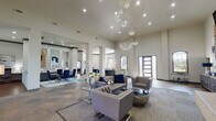 Located in Jersey Village, TX, this lobby and business center is a thoughtfully designed communal space that blends upscale aesthetics with practical functionality. The grand entrance immediately impresses with soaring ceilings adorned by cascading, globe-cluster crystal chandeliers that serve as a dramatic focal point throughout the open floor plan.

The main lounge area features rich slate tile flooring in warm earth tones, complemented by plush gray tufted sofas and armchairs accented with deep navy blue pillows and ottomans. A floor-to-ceiling stacked stone fireplace anchors one wall, topped by a mounted flat-screen TV, and flanked by tall windows dressed in sheer white curtains that invite abundant natural light and views of the surrounding greenery.

A dedicated business center area provides workstations with blue velvet chairs and built-in desk surfaces, while a separate consultation zone features a sleek reception-style desk. A glass-top dining table with bold royal blue velvet chairs sits against a mosaic tile accent wall with decorative mirrors, creating an elegant meeting or gathering spot. A rustic-modern credenza beneath a striking large-scale abstract blue artwork adds artistic character near the entry corridor.

Structural columns define the open layout, and recessed lighting throughout ensures a bright, welcoming ambiance suited to both professional and social interaction.