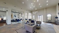 Located in Jersey Village, TX, this lobby and business center is a thoughtfully designed communal space that blends upscale aesthetics with practical functionality. The grand entrance immediately impresses with soaring ceilings adorned by cascading, globe-cluster crystal chandeliers that serve as a dramatic focal point throughout the open floor plan.

The main lounge area features rich slate tile flooring in warm earth tones, complemented by plush gray tufted sofas and armchairs accented with deep navy blue pillows and ottomans. A floor-to-ceiling stacked stone fireplace anchors one wall, topped by a mounted flat-screen TV, and flanked by tall windows dressed in sheer white curtains that invite abundant natural light and views of the surrounding greenery.

A dedicated business center area provides workstations with blue velvet chairs and built-in desk surfaces, while a separate consultation zone features a sleek reception-style desk. A glass-top dining table with bold royal blue velvet chairs sits against a mosaic tile accent wall with decorative mirrors, creating an elegant meeting or gathering spot. A rustic-modern credenza beneath a striking large-scale abstract blue artwork adds artistic character near the entry corridor.

Structural columns define the open layout, and recessed lighting throughout ensures a bright, welcoming ambiance suited to both professional and social interaction.