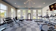 The fitness center at Ashton Brook in Louisville, Kentucky is a thoughtfully designed, single-floor facility built to support a well-rounded workout routine. The space features soaring ceilings accented with a bold teal-painted upper border, recessed lighting, and expansive floor-to-ceiling windows that flood the interior with natural light and provide views of the surrounding greenery and pool area. The checkerboard-patterned carpet tile flooring adds a clean, modern aesthetic throughout.

The cardio zone is well-stocked with multiple treadmills, elliptical machines, and stationary spinning bikes, complemented by a wall-mounted flat-screen television for entertainment during workouts. A full cable machine and multi-function strength station anchor the strength training area, supported by a tiered dumbbell rack with a full range of free weights and adjustable benches for versatile training. Stability balls and additional accessories round out the functional fitness offerings.

Large mirrors line key sections of the walls, visually expanding the space and aiding in form correction. Multiple glass doors provide direct outdoor access, enhancing ventilation and connectivity to the exterior grounds. The overall ambiance is bright, open, and motivating, making it a convenient and comprehensive amenity for residents of Ashton Brook.