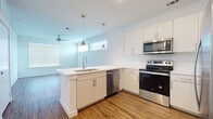 This single-floor residence showcases a clean, modern design with consistent finishes throughout. The open-concept kitchen is a standout feature, equipped with a full suite of stainless steel appliances including a refrigerator, electric range, over-the-range microwave, and dishwasher. White shaker-style cabinetry with brushed nickel hardware lines the walls, complemented by a white quartz countertop and a peninsula island with an undermount sink. Stylish pendant lights hang above the island, adding a contemporary touch. A textured tile backsplash and warm hardwood-style flooring complete the kitchen's polished look, which flows seamlessly into the open living area. The living space is bright and airy, benefiting from multiple windows with horizontal blinds and a ceiling fan overhead. The bathroom features a white quartz vanity with an undermount sink, modern chrome fixtures, a large mirror, and a glass-enclosed shower, all set on matching hardwood-style flooring. The bedrooms offer generous proportions, with large windows that invite abundant natural light. One bedroom features plush carpeting, while another showcases the same hardwood-style flooring found throughout the main living areas. Ceiling fans in the bedrooms add both comfort and a cohesive design element to this well-maintained, move-in-ready residence.