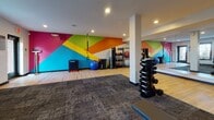 Located at 1000 Old Brook Rd in Charlottesville, Virginia, this single-floor fitness center is thoughtfully designed to support a well-rounded workout experience. The space is divided into distinct functional zones, each catering to different training styles and fitness goals.

The cardio area is equipped with stationary bikes — including an interactive Expresso cycling unit — as well as a rowing machine, all positioned near windows fitted with plantation-style blinds that allow natural light to filter in. A digital clock display and mounted screen keep users informed and engaged during their sessions.

The strength and free weights zone features a tiered dumbbell rack alongside a cable machine and additional resistance equipment. Large floor-to-ceiling mirrors line one wall, visually expanding the space and providing form-check capability during exercises. Structural columns are seamlessly integrated throughout.

A dedicated functional training area makes a bold visual statement with a striking, multi-colored geometric accent wall in shades of pink, orange, yellow, green, and blue. This zone includes TRX suspension trainers, kettlebells, medicine balls, a plyo box, and resistance bands, offering versatile training options.

The flooring transitions between hardwood and durable carpet tile, delineating workout zones while providing appropriate surfaces for each activity. Recessed lighting throughout ensures the space is well-lit, creating a clean, modern, and energizing atmosphere.