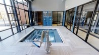 This indoor aquatic space features a compact heated pool or spa set within a sleek, modern facility designed with both functionality and accessibility in mind. The pool area is finished with light-colored large-format tile flooring arranged in a herringbone pattern, providing a clean and contemporary aesthetic throughout. Blue ceramic tile accents line the pool's interior and surrounding walls, adding a bold pop of color that complements the overall design palette.

A prominent ADA-compliant hydraulic pool lift is positioned at the pool's edge, ensuring accessible entry and exit for all users. Stainless steel handrails provide additional entry support along the pool's perimeter. The space is enclosed by floor-to-ceiling black-framed glass partitions and walls, which allow natural light to filter in while maintaining a sense of openness and transparency.

The elevated ceiling height adds a sense of volume and airiness to the space. Notices posted near the entrance indicate a maximum occupancy, suggesting this is a shared amenity facility. The overall ambiance is clean, bright, and purpose-built, reflecting a well-maintained and inclusive aquatic environment.