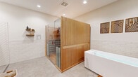 Located in Dacula, GA, this thoughtfully designed wellness room offers a comprehensive spa experience within a single, well-appointed space. The room is anchored by a striking cedar-clad infrared sauna enclosed with frameless glass doors, providing a warm, natural wood aesthetic that serves as the visual centerpiece. Adjacent to the sauna sits a freestanding Plunge cold therapy tub, enabling a seamless hot-cold contrast therapy routine.

The flooring throughout is finished in large-format hexagonal tiles in a soft gray tone, lending a clean, spa-like atmosphere. The lower portion of the walls features white stacked subway tile wainscoting, transitioning to smooth white-painted walls above, creating a crisp and modern aesthetic. Recessed lighting illuminates the space evenly, enhancing its open and airy feel.

A wall-mounted flat-screen television adds a layer of comfort and entertainment, while a teak slatted bench provides a practical spot to rest or prepare. Decorative wood wall art and dried botanical accents add warmth and personality to the otherwise minimalist design. Built-in shelving offers convenient storage for wellness accessories. A gray panel door provides a clean, modern entry point, completing this purpose-built retreat designed for relaxation and recovery.