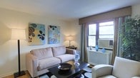This single-floor space showcases a thoughtfully designed living room that balances comfort and style. The living area is anchored by a plush, neutral-toned sofa positioned across from a sleek, dark coffee table, creating an inviting focal point for relaxation and conversation. A coordinating armchair adds additional seating while maintaining the room's cohesive aesthetic.

Natural light filters through a window dressed with elegant floor-length curtains, offering a view of greenery outside. The window also features a built-in air conditioning unit housed within a decorative white radiator cover, blending functionality with the room's clean design. A classic table lamp on a side console provides warm ambient lighting for the evenings.

The walls are adorned with two vibrant abstract art pieces in blue and green tones, adding personality and a pop of color against the neutral cream walls. Decorative blue glass vases and accent pieces on the windowsill and console contribute to a curated, polished look. A bold black-and-white area rug grounds the seating arrangement, adding visual contrast and texture to the overall space.