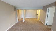 This single-floor space presents a spacious, open layout finished in soft, neutral tones throughout. The main living area is carpeted in warm beige, providing a comfortable and understated foundation that complements the light grey-blue walls. A textured popcorn ceiling adds classic character overhead, while white baseboards offer clean contrast along the perimeter.

A defining architectural feature of the main area is a slim structural column near the center of the space, subtly dividing the room while maintaining an open, connected feel. Two hallway openings branch off from the main living area — one to the left and one to the right — each leading to additional rooms illuminated by warm interior lighting. These passageways hint at a well-organized floor plan with dedicated spaces beyond the central living area.

The left hallway reveals at least one door, suggesting access to a bedroom or utility space, while the right opening leads to a warmly lit area accented by a small decorative plant, adding a touch of life to the interior. A wall-mounted HVAC vent on the left wall ensures year-round climate comfort. Overall, the space offers a clean, move-in-ready environment with a straightforward, functional layout.