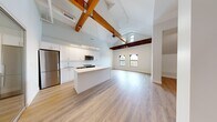 This thoughtfully designed one-bedroom, two-bathroom residence offers a generous and well-appointed single-floor layout. The standout feature is the dramatic open-concept kitchen and living area, where soaring vaulted ceilings with striking exposed wood beams create a sense of volume and loft-like character. Arched windows invite natural light to flood the space, complementing the light-toned hardwood flooring that runs throughout.

The kitchen is outfitted with stainless steel appliances, including a refrigerator, range, and built-in microwave, alongside crisp white cabinetry, a white quartz countertop island, and a clean subway tile backsplash. A dedicated hallway houses a stacked washer and dryer unit, adding in-unit laundry convenience.

The bedroom is a well-proportioned private retreat with neutral walls and warm wood flooring. A separate closet provides additional storage. The two bathrooms are finished with clean, contemporary fixtures — one features a glass-enclosed walk-in shower with white tile surround and beige marble-style tile flooring, while the second includes a full soaking tub with tiled surround. The overall ambiance is modern and refined, combining industrial architectural character with sleek, updated interiors.