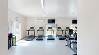 This fitness center offers a spacious, single-floor layout designed to accommodate a wide range of exercise activities. The interior is finished with clean white walls and light-toned flooring, creating a bright and motivating atmosphere throughout. Flush linear lighting fixtures line the ceiling, supplemented by abundant natural light streaming in through multiple large windows and glass doors that provide views of the surrounding greenery.

The cardio zone features a row of treadmills positioned along a windowed wall, alongside stationary bikes and other aerobic equipment, all facing mounted flat-screen televisions for an engaging workout experience. The strength training area is well-appointed with a multi-station cable machine, free weights and a dumbbell rack, weight benches, and additional resistance equipment. A wall-mounted mirror and workout signage support proper form and guidance.

A dedicated support area connects to the main workout floor and includes a wall-mounted water fountain with a bottle-filling station, a coat hook rail, and a flat-screen TV. A corridor leads to a separate room, offering additional utility to the facility. Climate control is provided via wall-mounted mini-split units. The overall design prioritizes functionality, openness, and comfort, making this a comprehensive and inviting fitness facility.