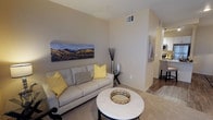 This single-floor residence offers a thoughtfully designed layout that balances comfort and functionality. The living area serves as the heart of the home, featuring neutral-toned walls that create a warm, inviting atmosphere. Plush carpeting underfoot adds a cozy feel, while the transition to hardwood-style flooring near the kitchen and dining area provides a clean contrast and visual interest.

The living room is well-proportioned, accommodating a generously sized sofa and a round marble-top coffee table that adds an elegant focal point. A large landscape canvas depicting a scenic mountain panorama adorns the wall, lending the space a nature-inspired character. Overhead, HVAC vents are neatly integrated into the ceiling, ensuring year-round comfort.

A hallway connects the living area to the rest of the home, with the kitchen visible in the background. The kitchen features light cabinetry and modern appliances, maintaining the overall clean and contemporary aesthetic. The open sightlines between rooms enhance the sense of spaciousness throughout.

Overall, the space presents a cohesive, modern design with neutral finishes, tasteful décor accents in gold and yellow tones, and a practical flow that makes it both livable and visually appealing.