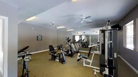 This fitness center offers a dedicated space for both cardio and strength training within a single, open-plan floor. The room features a practical, carpet-covered floor in a neutral tone that provides comfort underfoot during workouts. Multiple ceiling fans are evenly distributed throughout the space, ensuring steady airflow and a comfortable environment during exercise sessions. The walls are painted in a clean, neutral gray, contributing to a focused and motivating atmosphere. A large mirrored wall along one side of the room visually expands the space, allows users to monitor their form, and reflects natural light from the windows. The cardio section is well-stocked with upright and recumbent stationary bikes, elliptical trainers, and treadmills, catering to a range of cardiovascular fitness preferences. On the opposing side, a Paramount strength training machine provides options for targeted muscle conditioning. Informational signage is posted on the walls, offering guidance to users. Overhead fluorescent lighting supplements the natural light from windows, keeping the space bright throughout the day. Overall, the fitness center is thoughtfully arranged to maximize usable workout space while maintaining an open, accessible layout suitable for users of varying fitness levels.
