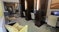 This single-floor business center is thoughtfully designed to support focused work in a refined, professional environment. The space is anchored by a series of semi-private workstations, each framed by tall, dark-paneled dividers that provide a sense of privacy while maintaining an open feel. Each station is equipped with a desktop computer and ergonomic white office chair, making them well-suited for individual work or quick business tasks. Integrated wall-mounted light fixtures between the panels cast a warm, ambient glow, contributing to a productive yet inviting atmosphere. The neutral-toned carpeted flooring adds a layer of acoustic comfort, reducing noise and enhancing concentration. Toward the front of the space, a casual lounge area features textured yellow-patterned accent chairs and a low coffee table, offering a relaxed spot for informal meetings or downtime between tasks. A full-length mirror and additional storage or display elements line the perimeter, adding depth and functionality to the layout. The overall design palette — rich dark wood tones contrasted with light upholstery and soft carpeting — creates a sophisticated, contemporary ambiance well-suited for a hotel business center or professional shared workspace.