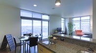 This single-floor residence showcases a thoughtfully designed open-concept layout that seamlessly connects the kitchen and living/dining areas. The kitchen is a standout feature, boasting expansive granite countertops with rich, multi-toned speckled patterning and warm wood cabinetry beneath. A pendant light fixture illuminates the kitchen island, which serves as a natural focal point and functional workspace. Floor-to-ceiling windows with horizontal blinds dominate the exterior walls, flooding the space with abundant natural light while framing impressive elevated city views that stretch across the urban skyline. The dining area, positioned directly adjacent to the windows, features a glass-top table paired with sleek, dark upholstered chairs, creating an elegant setting with a scenic backdrop. Recessed ceiling lighting complements the natural light throughout. A media console with a flat-screen television anchors the living area, while decorative bamboo plants and tasteful accents add warmth and organic texture. The overall ambiance is modern and refined, combining contemporary finishes with an airy, light-filled atmosphere characteristic of elevated urban living.