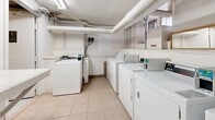 Located at 1245 Rue St-Marc in Montréal's vibrant H3H neighbourhood, this building amenity space offers residents two practical and functional areas designed to support everyday living. The laundry room is well-appointed with multiple commercial-grade top-loading washing machines and front-loading dryers arranged in efficient rows, all equipped with digital payment controls. Light tile flooring, white walls, and overhead fluorescent lighting create a clean, bright environment. A built-in folding counter runs along one wall, providing a convenient surface for sorting and folding laundry. Informational signage and instructions are posted throughout, and a stainless steel waste bin adds a tidy finishing touch. Exposed overhead piping and ventilation ducts contribute to the room's utilitarian character. Adjacent to the laundry area, the indoor bicycle storage room offers secure, dedicated space for residents' bikes, featuring mounted floor-level bike racks capable of accommodating multiple bicycles. The room is finished with a smooth blue-painted concrete floor and white walls, lit by strip fluorescent lighting. Several lockable metal-grate storage lockers line one wall, providing additional secure storage options. A fire extinguisher is conveniently mounted for safety. Together, these amenities reflect a well-maintained, resident-focused building.