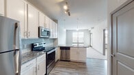 This single-floor residence offers a thoughtfully designed open-concept layout that seamlessly connects the kitchen and living areas. The kitchen is a standout feature, showcasing crisp white cabinetry with brushed nickel hardware, granite countertops, a light blue subway tile backsplash, and a full suite of stainless steel appliances including a refrigerator, electric range, microwave, and dishwasher. A peninsula with additional counter space and a sink anchors the kitchen, while pendant lighting and track lighting illuminate the space. Wood-look flooring runs throughout the main living areas, adding warmth and continuity.

The bedrooms are carpeted and filled with natural light from large windows with exterior views. A ceiling fan is featured in one bedroom, enhancing comfort. The primary bathroom is spacious and well-appointed, featuring wood-look tile flooring, a walk-in glass-enclosed shower, a granite-topped vanity, and ample storage. A generously sized walk-in closet with wire shelving provides excellent storage capacity.

The overall aesthetic is clean and contemporary, with a neutral color palette of whites and soft grays creating a bright, welcoming ambiance throughout the space.