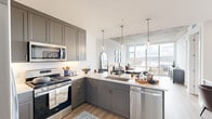 This thoughtfully designed one-bedroom, one-bathroom residence offers a seamless open-concept layout highlighted by striking architectural details and stunning waterfront views. The living and kitchen areas flow together effortlessly, anchored by dramatic floor-to-ceiling windows that frame expansive views of the river and surrounding landscape, flooding the space with natural light. Exposed concrete ceiling panels add an industrial-modern character that contrasts beautifully with the clean white walls and warm hardwood-style flooring throughout.

The kitchen is well-appointed with gray shaker cabinetry, white quartz countertops, a subway tile backsplash, and a full suite of stainless steel appliances including a gas range, built-in microwave, dishwasher, and French door refrigerator. A central island with pendant lighting provides additional prep space and casual seating. The living area benefits directly from the panoramic window wall, creating a bright and airy atmosphere ideal for relaxing or entertaining.

The bedroom continues the floor-to-ceiling window theme, offering the same sweeping outdoor views and an exposed concrete ceiling. It connects to a well-organized walk-in closet with open shelving and hanging rods. The bathroom features gray cabinetry, a dual-sink vanity with quartz counters, and in-unit laundry conveniently stacked nearby.