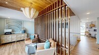 Located in Little River, South Carolina, this thoughtfully designed single-story amenity space offers a compelling blend of coastal aesthetics and functional programming. The interior is anchored by a striking lounge area defined by a dramatic floor-to-ceiling slatted wood partition accented with nautical rope detailing, creating a visual focal point that echoes the surrounding coastal environment. Warm wood plank flooring, a natural wood-paneled ceiling, and a woven shell chandelier lend the space an inviting, resort-like ambiance. Soft blue-green walls and teal accent pillows reinforce the coastal palette throughout.

The fitness center is generously equipped with a full complement of strength and cardio machines, including cable systems, treadmills, ellipticals, a rowing machine, and free weights, all reflected by floor-to-ceiling mirrors. Large windows and direct outdoor access flood the gym with natural light and views of the pool area beyond.

A dedicated co-working zone features a long built-in desk with multiple workstations, ample cabinet storage below, and a gallery-style wall of framed artwork above. Adjacent to this workspace is a well-appointed kitchenette with slate-blue cabinetry, a undermount sink, and a Starbucks coffee station, providing a convenient refresh point. Together, these areas create a versatile, amenity-rich environment suited for both active and productive use.