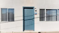 This single-story unit presents a straightforward and functional layout designed for everyday living. The exterior is characterized by a light-colored stucco façade, accented by a bold teal six-panel entry door with a wall-mounted light fixture alongside it, offering a welcoming and easily identifiable entrance.

The kitchen is a central feature of the space, designed with practicality in mind. Warm honey-toned wood cabinetry lines both the upper and lower walls, providing ample storage throughout. The countertops feature a granite-style surface in earthy brown tones, offering durable workspace on both sides of the kitchen. A double-basin stainless steel sink sits beneath upper cabinets on one side, while the opposite wall accommodates a full suite of black appliances, including a gas range with multiple burners, a built-in range hood above, and a full-sized refrigerator. Dark wood-style flooring runs throughout the kitchen, adding warmth and contrast to the white walls. Overhead, a flush-mount ceiling light provides bright, even illumination across the space.