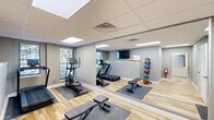 Located at 36 S Sixth St in Easton, Pennsylvania, this fitness facility offers a thoughtfully designed exercise space and a modern restroom within a single, efficiently organized floor. The exercise room features light hardwood flooring that adds warmth to the neutral gray walls, while a drop ceiling with flush LED panel lighting and a skylight ensures a bright, energizing atmosphere. Floor-to-ceiling mirrors along one wall visually expand the space and support proper form during workouts. The room is equipped with a treadmill, elliptical, adjustable weight bench, medicine ball rack, dumbbell rack with a full range of free weights, and kettlebells — providing a comprehensive range of cardiovascular and strength training options. A wall-mounted flat-screen TV with a small counter area adds a functional informational hub to the space. Natural light filters in through a window, further enhancing the open feel of the room. The adjacent restroom is ADA-accessible and finished with striking dark slate tile flooring and warm vertical wood slat wall paneling, creating a refined, spa-like contrast. It features a modern pedestal sink, an illuminated frameless mirror, and clean white millwork throughout. Together, these two spaces form a complete, polished fitness facility suited for focused, full-body training.