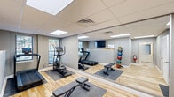 Located at 36 S Sixth St in Easton, Pennsylvania, this fitness facility offers a thoughtfully designed exercise space and a modern restroom within a single, efficiently organized floor. The exercise room features light hardwood flooring that adds warmth to the neutral gray walls, while a drop ceiling with flush LED panel lighting and a skylight ensures a bright, energizing atmosphere. Floor-to-ceiling mirrors along one wall visually expand the space and support proper form during workouts. The room is equipped with a treadmill, elliptical, adjustable weight bench, medicine ball rack, dumbbell rack with a full range of free weights, and kettlebells — providing a comprehensive range of cardiovascular and strength training options. A wall-mounted flat-screen TV with a small counter area adds a functional informational hub to the space. Natural light filters in through a window, further enhancing the open feel of the room. The adjacent restroom is ADA-accessible and finished with striking dark slate tile flooring and warm vertical wood slat wall paneling, creating a refined, spa-like contrast. It features a modern pedestal sink, an illuminated frameless mirror, and clean white millwork throughout. Together, these two spaces form a complete, polished fitness facility suited for focused, full-body training.