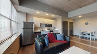 Located at 901 S National Ave in Springfield, Missouri, this single-floor unit showcases a contemporary industrial aesthetic defined by exposed concrete ceilings, warm wood-style flooring, and neutral gray tones throughout. The open-concept living and kitchen area forms the heart of the space, where generous windows allow abundant natural light and provide views of the surrounding urban streetscape. The elevated ceiling height adds a sense of openness to the living area, which comfortably accommodates a seating arrangement centered around a coffee table and wall-mounted television.

The kitchen is efficiently designed with sleek light-toned cabinetry, white countertops, and a full suite of stainless steel appliances, including a refrigerator, range, microwave, and dishwasher. Recessed lighting ensures the kitchen remains bright and functional.

The bedroom continues the unit's clean, modern design with the same wood-style flooring, soft gray walls, and a well-positioned window that invites natural light. The room offers space for a bed and a dedicated desk area, making it well-suited for both rest and work.

The bathroom is finished with coordinating flooring and neutral walls, featuring a shower and standard fixtures with brushed nickel hardware accents. Overall, the unit presents a cohesive, urban-modern atmosphere.