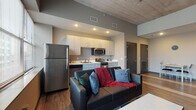 Located at 901 S National Ave in Springfield, Missouri, this single-floor unit showcases a contemporary industrial aesthetic defined by exposed concrete ceilings, warm wood-style flooring, and neutral gray tones throughout. The open-concept living and kitchen area forms the heart of the space, where generous windows allow abundant natural light and provide views of the surrounding urban streetscape. The elevated ceiling height adds a sense of openness to the living area, which comfortably accommodates a seating arrangement centered around a coffee table and wall-mounted television.

The kitchen is efficiently designed with sleek light-toned cabinetry, white countertops, and a full suite of stainless steel appliances, including a refrigerator, range, microwave, and dishwasher. Recessed lighting ensures the kitchen remains bright and functional.

The bedroom continues the unit's clean, modern design with the same wood-style flooring, soft gray walls, and a well-positioned window that invites natural light. The room offers space for a bed and a dedicated desk area, making it well-suited for both rest and work.

The bathroom is finished with coordinating flooring and neutral walls, featuring a shower and standard fixtures with brushed nickel hardware accents. Overall, the unit presents a cohesive, urban-modern atmosphere.