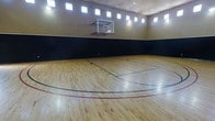 This dedicated indoor sports facility is centered around a full-size basketball court with a pristine, regulation-grade hardwood floor. The polished wood surface features clearly marked court lines in multiple colors, including three-point arcs and key areas, providing a professional playing experience. A wall-mounted glass backboard and hoop is positioned at one end of the court, meeting standard play requirements.

The court is enclosed by walls finished in a two-tone design — a deep charcoal lower panel that serves as a protective barrier along the lower half of the walls, transitioning to a warm tan upper section that extends to the ceiling. This design helps define the playing zone while offering a clean, finished aesthetic.

One of the standout architectural features is the row of clerestory windows that runs along the upper perimeter of the space. These high windows invite generous amounts of natural light into the interior, reducing reliance on artificial lighting during daytime use and creating an open, airy atmosphere despite being fully enclosed. The soaring ceiling height further enhances the sense of volume, ensuring ample vertical clearance for gameplay. Supplemental overhead lighting ensures the space remains well-illuminated at all hours. Overall, this court offers a polished, purpose-built environment ideal for recreational or competitive basketball and other indoor sports activities.