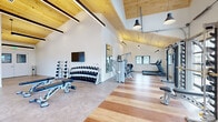 Located in Colorado Springs, CO, this dedicated fitness center offers a spacious and thoughtfully designed workout facility. The space is immediately defined by its striking vaulted ceiling clad in warm natural wood planking, accented by dark exposed beams and modern linear pendant lighting that creates a bright, energizing atmosphere throughout.

The flooring transitions seamlessly between polished concrete and rich hardwood planks, delineating functional workout zones. Large glass-panel garage-style doors along the perimeter flood the interior with natural light and offer open views of the surrounding Colorado landscape, while additional windows provide ample ventilation and daylight.

The strength training area features a full rack of dumbbells, adjustable workout benches, a cable machine system, and a multi-function Hoist weight machine, complemented by wall-mounted mirrors that allow users to monitor their form. The cardio zone is lined with treadmills and a stair climber, positioned to take advantage of the natural light from the expansive windows and garage doors.

A wall-mounted flat-screen television adds a media element to the training environment. The clean white walls contrast beautifully against the warm wooden ceiling, giving the space a modern yet welcoming aesthetic. The overall design balances functionality with architectural character, making this fitness center a standout amenity.