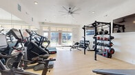 Located on Dry Creek Drive in Longmont, Colorado, this fitness center delivers a comprehensive workout environment within a thoughtfully designed single-room layout. The space features light hardwood-style flooring, crisp white walls, and elevated ceilings fitted with recessed lighting and a large ceiling fan, creating a clean and energizing atmosphere throughout.

Large sliding glass doors along one wall flood the room with natural light and provide direct views of the surrounding outdoor area, enhancing the open feel of the space. A full wall of mirrors on one side visually expands the room and allows for form monitoring during workouts.

The cardio section is well-appointed with multiple treadmills, stair climbers, and a rowing machine. For strength training, the space includes a robust power rack system with pull-up bars, a full dumbbell rack spanning a wide weight range, kettlebells, medicine balls, and weight plates. Additional equipment includes a functional trainer cable machine, a spin bike, a stability ball, an adjustable weight bench, and a plyo box. A wall-mounted flat-screen TV is also present, providing entertainment during workouts.

The space is efficiently organized to maximize usability, offering a well-rounded facility suitable for cardio, strength, and functional fitness training.