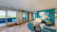 Located at 9200 Bruckhaus St in Raleigh, NC, this thoughtfully designed community lounge space offers a welcoming and energetic atmosphere defined by a carefully curated modern aesthetic. The open-plan layout flows seamlessly across light-toned tile flooring, creating a sense of spaciousness throughout.

A striking teal accent wall serves as the visual centerpiece of the lounge area, adorned with decorative metallic disc wall art and complemented by a bold sculptural pendant light that draws the eye upward. Teal upholstered seating, accented with chartreuse throw pillows, is arranged around low coffee tables atop a patterned area rug, offering comfortable conversation zones.

Toward the center of the space, high-top tables and chairs provide a more casual dining or work setting, while a dedicated nook with booth-style banquette seating, wood-topped tables, and matching pendant fixtures offers a more intimate gathering spot, ideal for collaborative use.

Large windows dressed in vibrant multicolored striped drapery flood the interior with natural light and frame views of the surrounding Raleigh landscape. Recessed ceiling lighting ensures the space is well-lit throughout. Built-in cabinetry provides practical storage, and curated artwork enhances the walls. Overall, the space strikes a balance between lively design and functional comfort.