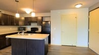 This single-floor residence offers a thoughtfully designed open-concept layout that seamlessly connects the kitchen and living areas, creating a functional and inviting atmosphere. The kitchen is a standout feature, showcasing rich dark cabinetry that provides ample storage, complemented by a peninsula with a light-toned countertop that doubles as a prep and casual dining space. A stainless steel sink is integrated into the peninsula, adding convenience. Black appliances, including a refrigerator, range, and microwave, lend a sleek, cohesive look to the culinary space. Pendant lighting above the kitchen area adds warmth and a touch of style, enhancing the overall ambiance. The adjoining open area is finished with durable hardwood-style flooring that runs throughout, offering both aesthetic appeal and easy maintenance. Neutral-toned walls keep the space feeling bright and versatile, ready to accommodate a variety of interior styles. A full-panel interior door leads to an adjacent room, reinforcing the practical flow of the floor plan. The layout is efficient and well-organized, making it an ideal space for comfortable everyday living.