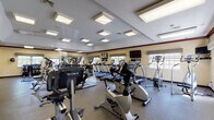 The clubhouse at Hillcrest Village in Holbrook, New York is a single-story community facility designed to support both active and social lifestyles. The well-equipped fitness center is a standout feature, housing a wide array of cardio machines including treadmills, ellipticals, stationary bikes, and stair climbers, predominantly from the True Fitness brand. Large wall mirrors amplify the sense of space, while multiple flat-screen TVs and natural light from several windows create an energizing workout environment. Durable rubber flooring and recessed panel ceiling lights complete the practical, clean aesthetic. A dedicated free weights area includes a dumbbell rack, adjustable bench, and additional mirrors.

Adjacent to the fitness center is a comfortable recreation lounge featuring a full-size pool table, leather sofas, and a dining table with blue upholstered chairs — ideal for casual socializing. A compact kitchenette with wood cabinetry, a microwave, sink, and mini-fridge provides added convenience.

The leasing office area includes a welcoming waiting nook with seating and framed floor plan displays, alongside a private office furnished with a wood desk, computer workstation, filing cabinets, and community artwork. Tile flooring runs throughout the common areas, tying the facility together with a warm, consistent finish.