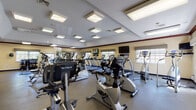 The clubhouse at Hillcrest Village in Holbrook, New York is a single-story community facility designed to support both active and social lifestyles. The well-equipped fitness center is a standout feature, housing a wide array of cardio machines including treadmills, ellipticals, stationary bikes, and stair climbers, predominantly from the True Fitness brand. Large wall mirrors amplify the sense of space, while multiple flat-screen TVs and natural light from several windows create an energizing workout environment. Durable rubber flooring and recessed panel ceiling lights complete the practical, clean aesthetic. A dedicated free weights area includes a dumbbell rack, adjustable bench, and additional mirrors.

Adjacent to the fitness center is a comfortable recreation lounge featuring a full-size pool table, leather sofas, and a dining table with blue upholstered chairs — ideal for casual socializing. A compact kitchenette with wood cabinetry, a microwave, sink, and mini-fridge provides added convenience.

The leasing office area includes a welcoming waiting nook with seating and framed floor plan displays, alongside a private office furnished with a wood desk, computer workstation, filing cabinets, and community artwork. Tile flooring runs throughout the common areas, tying the facility together with a warm, consistent finish.