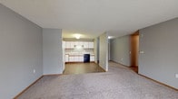 This single-floor unit offers a practical, open-concept design with clearly defined living and kitchen zones. The living area is finished with neutral gray walls, plush wall-to-wall carpeting, and a textured ceiling. A large sliding glass door fitted with vertical blinds fills the space with natural light and provides outdoor views, making the area feel open and inviting.

The kitchen is efficiently laid out in an L-shaped configuration and transitions from the living area via tile flooring. It features white cabinetry with wood-tone trim offering generous upper and lower storage, light-colored laminate countertops, a stainless steel double-basin sink with an under-cabinet light fixture, a black dishwasher, and a white electric range with a ventilation hood. The combination of white cabinets and neutral walls creates a clean, functional cooking environment.

The bathroom includes tile flooring consistent with the kitchen, a white soaking tub and shower combination, a vanity with white cabinetry and wood-tone accents, a large mirror, and a lighted fixture above the sink area. The overall unit maintains a cohesive, neutral color palette of gray and white throughout, contributing to a clean and understated aesthetic.