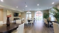 This single-floor space is thoughtfully designed to serve as a communal gathering and dining area, combining functionality with a refined aesthetic. The room is anchored by rich hardwood flooring that runs throughout, lending warmth and continuity to the space. Multiple dark-toned dining tables are paired with tufted white upholstered chairs featuring nailhead trim detailing, creating an elegant yet inviting atmosphere.

Natural light fills the interior through a series of tall arched windows and glass French doors, offering views of the surrounding outdoor greenery. Sheer white curtains frame the windows, softening the light while maintaining an airy, open feel. Recessed ceiling lighting provides additional illumination throughout the space.

On one end of the room, a wall-mounted flat-screen television rests above a dark console table, creating a media-friendly lounge area that complements the dining setup. Decorative elements, including potted tropical plants and wall art, add color and texture to the neutral cream-toned walls. The overall ambiance is clean, modern, and welcoming, making this space well-suited for social gatherings, dining, or casual meetings.
