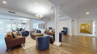 Located at 187 Paterson Ave in Midland Park, NJ, this single-floor community space blends classic architectural detail with a warm, functional design. The grand entrance features a polished tile foyer framed by a reception desk, decorative columns, large glass entry doors, and a chandelier that sets an inviting tone from the moment of arrival.

The expansive main lounge is anchored by tall white columns with ornate capitals, gleaming hardwood floors, and elegant chandeliers that cast a soft, welcoming glow. Comfortable seating arrangements—including tufted navy leather wingback chairs, club chairs, and upholstered sofas—are thoughtfully arranged throughout, creating intimate gathering areas within the open layout. Wainscoting and crown molding add a refined architectural touch to the crisp white walls.

A built-in white bookcase, fully stocked with a wide selection of reading materials, lines one wall of the lounge and extends into a sunlit reading alcove. This area features French doors that open to an outdoor patio, flooding the space with natural light and garden views. A warm wood upright piano nestled nearby adds a cultural focal point to the room.

A dedicated computer room provides a quiet, carpeted workspace equipped with workstations, a printer, and a community bulletin board, offering residents a practical and organized area for personal and administrative needs.