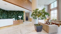 Located at 900 Pacific Street in Stamford, Connecticut, Opus presents a meticulously designed ground-floor amenity level that blends luxury finishes with a warm, nature-inspired aesthetic. The grand lobby greets visitors with soaring double-height ceilings, large-format white marble-effect tile flooring, and dramatic floor-to-ceiling windows that flood the space with natural light and offer views of the surrounding greenery. A living green wall behind the reception desk serves as a striking focal point, complemented by lush potted trees throughout that reinforce the biophilic design philosophy.

A central lounge area features an impressive full-height marble fireplace surround, plush white seating, and rich wood paneling that adds warmth and texture. Adjacent to the lobby, a spacious social lounge offers warm hardwood-style flooring, a mix of intimate seating arrangements, a bar counter, and a shimmering gold accent wall, all bathed in natural light from expansive windows. Gold-accented railings and illuminated stepped platforms add architectural dimension throughout the transition spaces.

The amenity corridor leads to dedicated conference and meeting areas, branded with the Opus identity and furnished with refined, contemporary pieces. Recessed and cove lighting throughout creates a consistent, polished ambiance across every zone of this distinguished space.