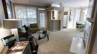 This single-story space offers a comfortable, open-concept layout that seamlessly connects the living area to the kitchen. The living room is carpeted and bathed in natural light, thanks to large windows and a glass-paneled exterior door that frame views of lush greenery outside. Crown molding adds a refined architectural detail throughout, complementing the warm, neutral taupe wall tones. The living area comfortably accommodates a dark leather sofa, accent chair in green upholstery, and a distinctive glass-topped coffee table with a sculptural base, contributing to a contemporary aesthetic. A raised pass-through counter separates the living space from the kitchen, which is visible in the background and features warm wood cabinetry and full-sized appliances, including a refrigerator. Beyond the kitchen, a hallway leads further into the home, suggesting additional rooms. The overall ambiance is warm and inviting, combining practical layout design with tasteful finishes that create a cohesive and functional living environment throughout the single floor.