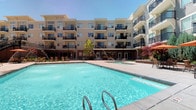 This residential community is centered around a spacious outdoor courtyard that serves as a social and recreational hub. The highlight of the exterior is a generously sized swimming pool with crystal-clear turquoise water, framed by a clean tile deck and equipped with entry handrails for easy access. Poolside lounge chairs and red seating areas are arranged along the perimeter, offering comfortable spaces to relax in the sun.

The surrounding multi-story building features a neutral stucco façade with large windows and private balconies fitted with dark metal railings, providing residents with outdoor space and views of the courtyard below. The architectural design blends contemporary and traditional elements, with structured massing and clean lines throughout.

Mature ornamental trees, including a striking red Japanese maple, add natural color and softness to the hardscaped courtyard. Ground-level commercial or amenity spaces with glass storefronts line the base of the building, while classic-style lamp posts contribute to the overall polished aesthetic. The open-air design allows for abundant natural light throughout the day.