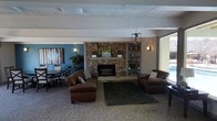 This single-floor space offers a warm and inviting atmosphere designed for comfortable gathering and relaxation. The centerpiece of the main lounge area is a striking floor-to-ceiling stacked stone fireplace, flanked by built-in shelving units that provide both display space and architectural symmetry. A pair of rich brown leather armchairs and a plush area rug are arranged in front of the fireplace, creating an intimate seating arrangement ideal for conversation or quiet rest. The room features a combination of textured ceiling panels and exposed beams, adding dimension and character overhead. A modern pendant chandelier provides ambient lighting, complementing the wall-mounted sconces that contribute to the warm glow throughout the space. Adjacent to the lounge, a dining area is defined by a bold teal accent wall adorned with decorative artwork, adding a pop of color and visual contrast to the neutral tones of the main room. Dark wood dining chairs and tables offer a functional yet stylish space for meals or meetings. Natural light filters in through a large sliding glass door, connecting the interior to the outdoors and enhancing the open, airy feel of the space. Overall, the design balances comfort, functionality, and thoughtful décor throughout.