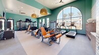 Located at 2935 W Lancaster Blvd in Lancaster, California, this fitness center offers a comprehensive workout facility spread across a single level. The main exercise room is defined by its striking teal-painted walls, elevated ceilings, and an abundance of natural light streaming through large arched and floor-to-ceiling windows that frame views of palm trees and the surrounding community. Stylish woven pendant light fixtures and ceiling fans add a welcoming, modern aesthetic to the space. The interior is outfitted with a wide array of cardio and strength-training equipment, including treadmills, ellipticals, a stationary bike with an interactive display, cable machines, multi-functional strength stations, a full dumbbell rack, and orange-accented resistance machines. Wall-to-wall mirrors enhance the sense of depth, while a wall-mounted flat-screen TV keeps members entertained during workouts. A decorative tilework accent wall adds a distinctive design touch. Connecting hallways lead to a semi-enclosed outdoor turf training area, which features a power rack with weight plates, a bench, and battle ropes beneath a dramatic arched opening — offering an open-air training experience with views of the community grounds. Together, these spaces provide a versatile and fully equipped environment for a wide range of fitness activities.