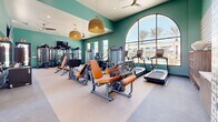 Located at 2935 W Lancaster Blvd in Lancaster, California, this fitness center offers a comprehensive workout facility spread across a single level. The main exercise room is defined by its striking teal-painted walls, elevated ceilings, and an abundance of natural light streaming through large arched and floor-to-ceiling windows that frame views of palm trees and the surrounding community. Stylish woven pendant light fixtures and ceiling fans add a welcoming, modern aesthetic to the space. The interior is outfitted with a wide array of cardio and strength-training equipment, including treadmills, ellipticals, a stationary bike with an interactive display, cable machines, multi-functional strength stations, a full dumbbell rack, and orange-accented resistance machines. Wall-to-wall mirrors enhance the sense of depth, while a wall-mounted flat-screen TV keeps members entertained during workouts. A decorative tilework accent wall adds a distinctive design touch. Connecting hallways lead to a semi-enclosed outdoor turf training area, which features a power rack with weight plates, a bench, and battle ropes beneath a dramatic arched opening — offering an open-air training experience with views of the community grounds. Together, these spaces provide a versatile and fully equipped environment for a wide range of fitness activities.