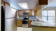 This single-floor space showcases a thoughtfully designed kitchen that serves as a central feature of the home. The kitchen is outfitted with a full suite of stainless steel appliances, including a french-door refrigerator, an electric range with oven, and an over-the-range microwave, providing a cohesive and modern aesthetic. Warm honey-toned wood cabinetry lines the walls, offering generous upper and lower storage space, while a decorative mosaic tile backsplash adds visual texture and character between the countertops and cabinets. The countertops display a granite finish with soft earth-tone veining, complementing the warm cabinetry tones throughout. A double stainless steel sink is conveniently positioned near a window, allowing natural light to filter into the workspace. The kitchen also features a peninsula-style counter extension, offering additional prep space and a natural division between the kitchen and adjacent areas. Light ceramic tile flooring runs throughout, contributing to a clean, easy-to-maintain environment. A recessed ceiling panel with integrated lighting provides even illumination across the entire kitchen workspace.
